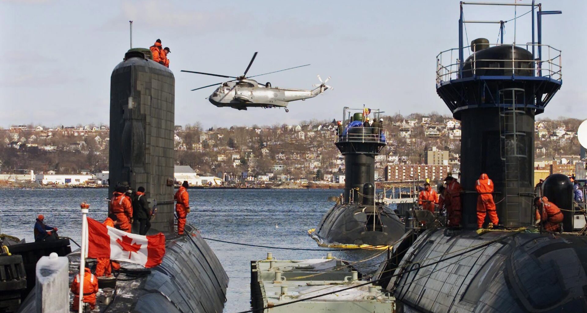 Three submarines docked side by side, crew in orange suits atop them. A helicopter hovers above, a Canadian flag visible. Coastal view of Halifax in the background.