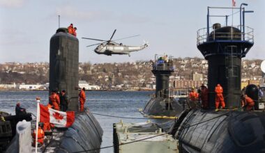 Three submarines docked side by side, crew in orange suits atop them. A helicopter hovers above, a Canadian flag visible. Coastal view of Halifax in the background.