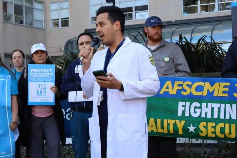 A man in a white lab coat speaks into a microphone at an outdoor rally, with others holding signs that reference patient care and union support in the background.