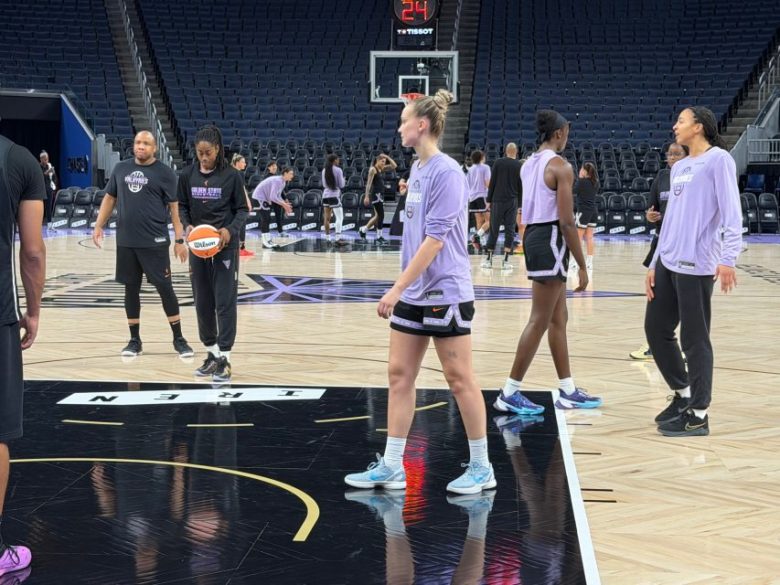 Basketball players in purple practice shirts warm up on an indoor court, with coaches and other players in the background. The arena seats are mostly empty.
