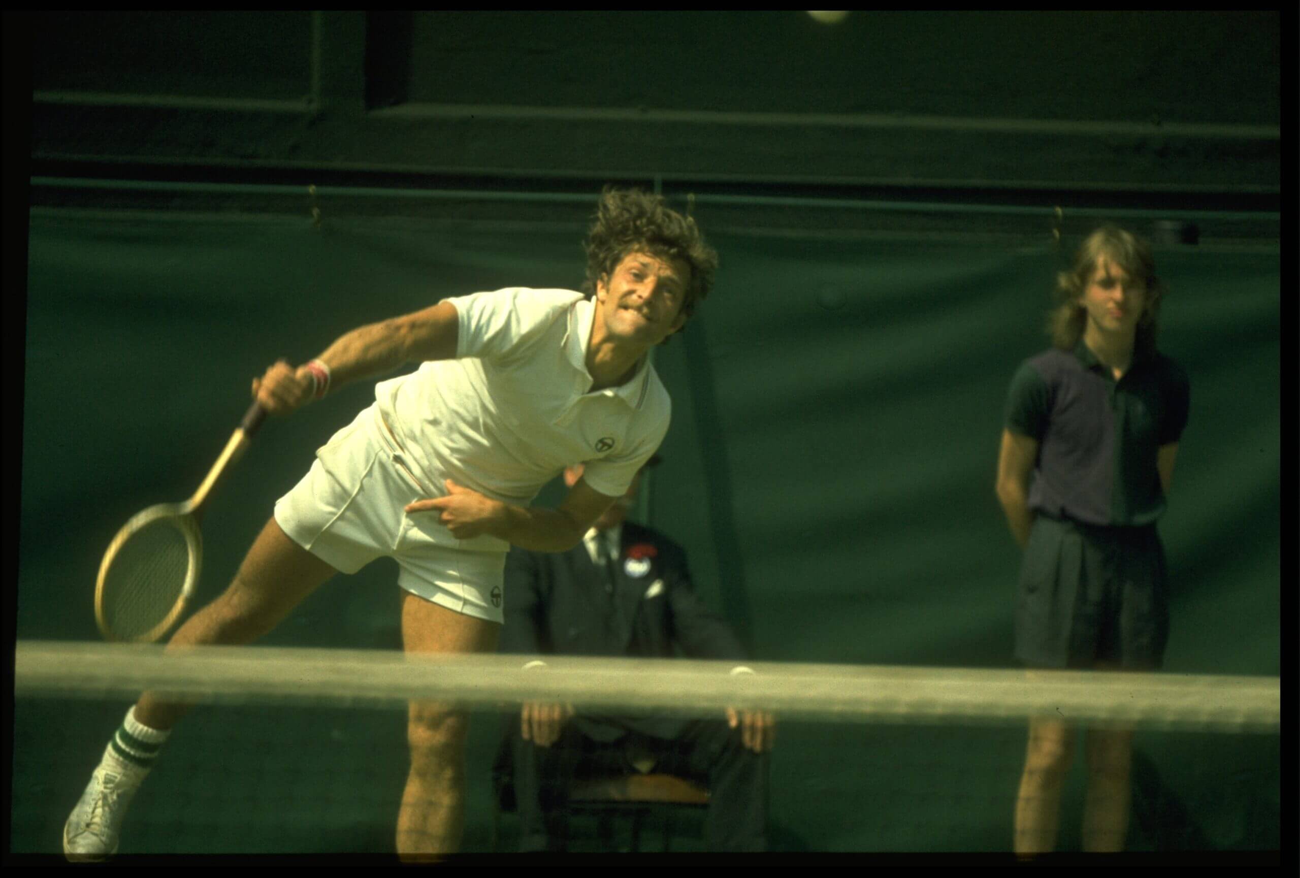 Jan Kodeš follows through on a serve wearing an all-white tennis outfit and using a wooden racket, as a ball kid watches on.