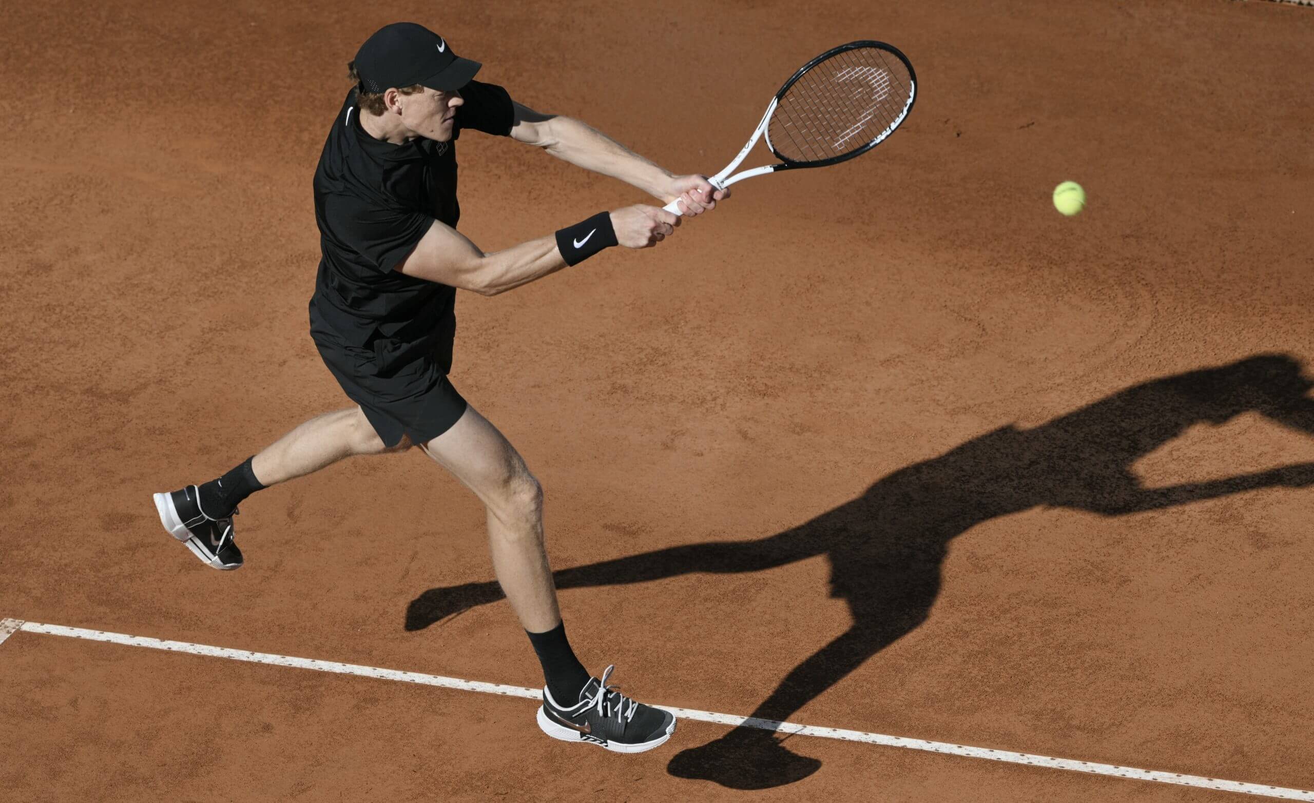 Jannik Sinner hits a two-handed backhand in mid-air on a clay tennis court wearing an all-black outfit.