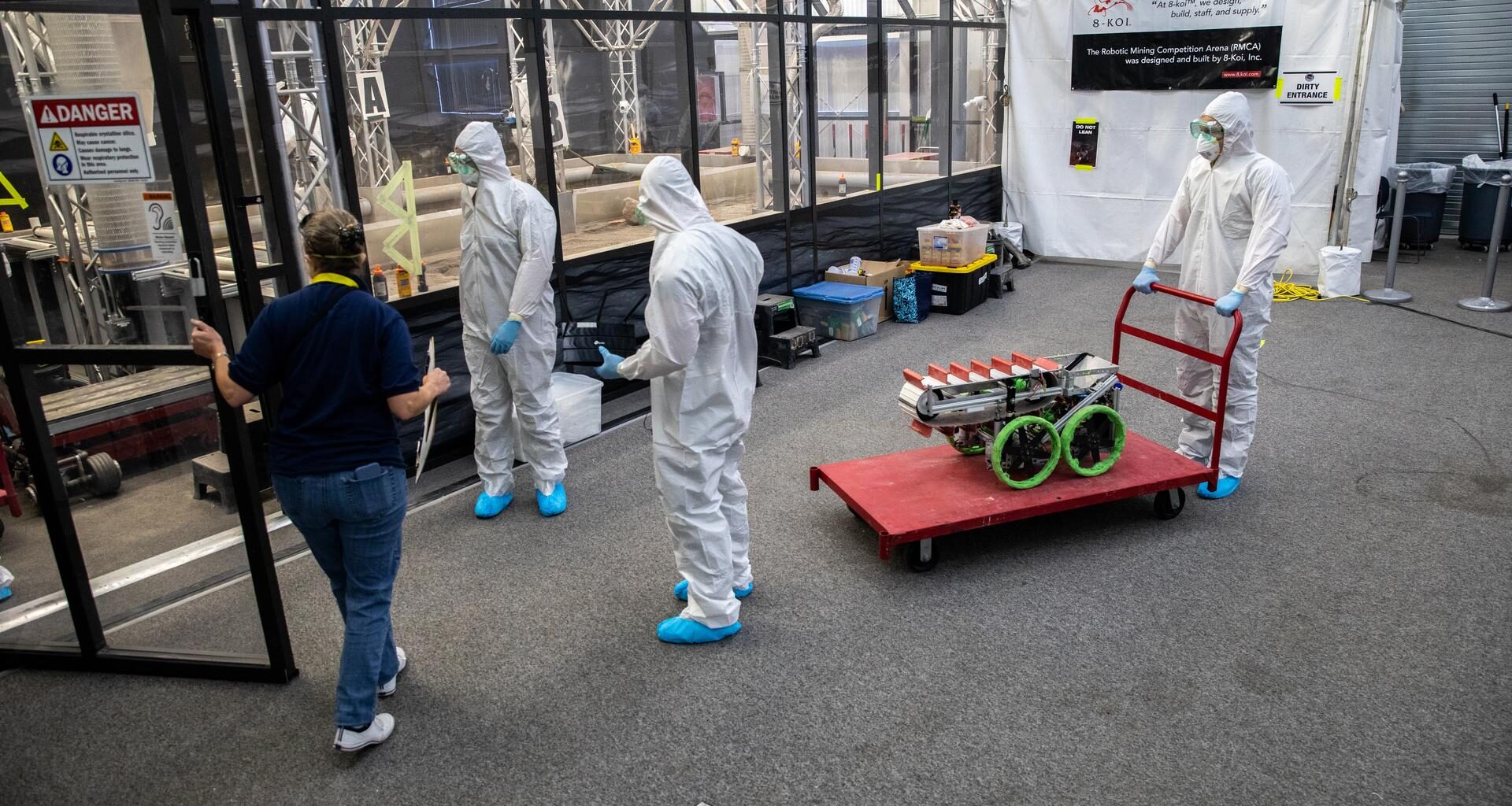 Image shows three people in white protective suits inside a room with another person wearing a blue shirt and blue jeans at NASA's Kennedy Space Center in Florida in 2022. Around them is a red cart with a silver structure and green looking wheels. Photo credit: NASA/Kim Shiflett