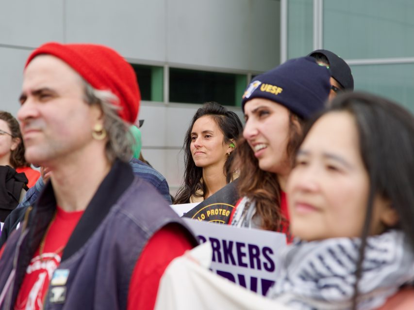 A group of people stand together at a protest or rally, some holding signs and expressing their views, while the risk of arrest looms in the background near a tall building.