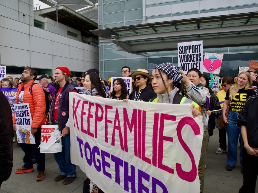A group of people hold signs at a protest, including one large banner reading "KEEP FAMILIES TOGETHER," demanding an end to unjust arrest practices outside a modern building.