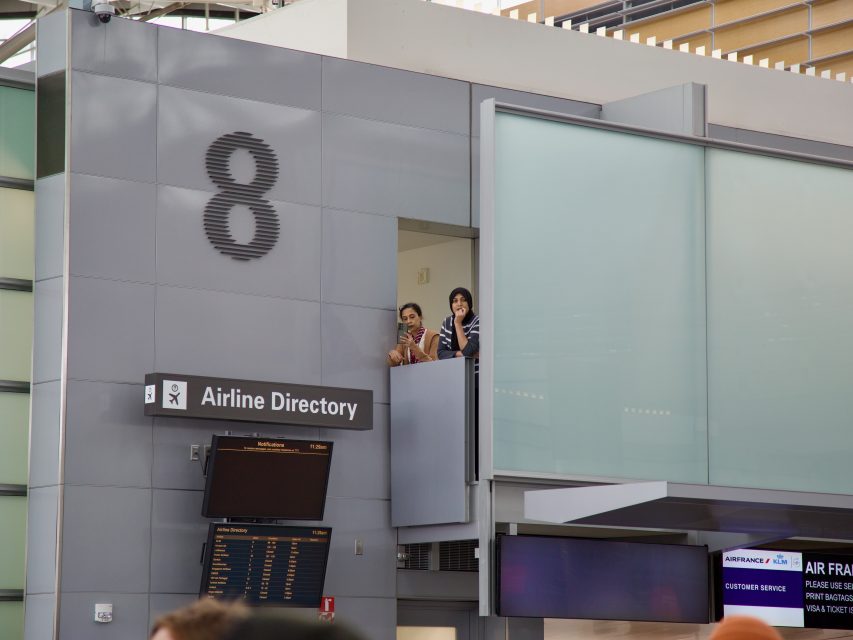Two people stand on a balcony above an Airline Directory sign and flight information board at an airport terminal marked with the number 8, unaware of a nearby arrest unfolding below.