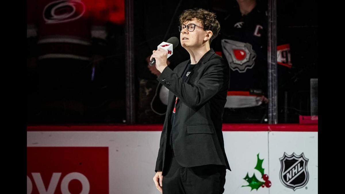 Mason Greer sings the Canadian national anthem ahead of the Carolina Hurricanes’ game against the Edmonton Oilers on Nov. 22, 2023. Mason Greer, a white man with curly brown hair, glasses and a black suit jacket with a Carolina Hurricanes jersey underneath, holds a Canes-branded microphone while he sings the national anthem.