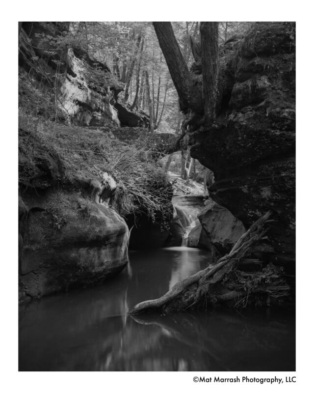 Black and white photo of a serene creek flowing between high rocky cliffs and boulders, surrounded by trees and thick vegetation, with reflections on the calm water.