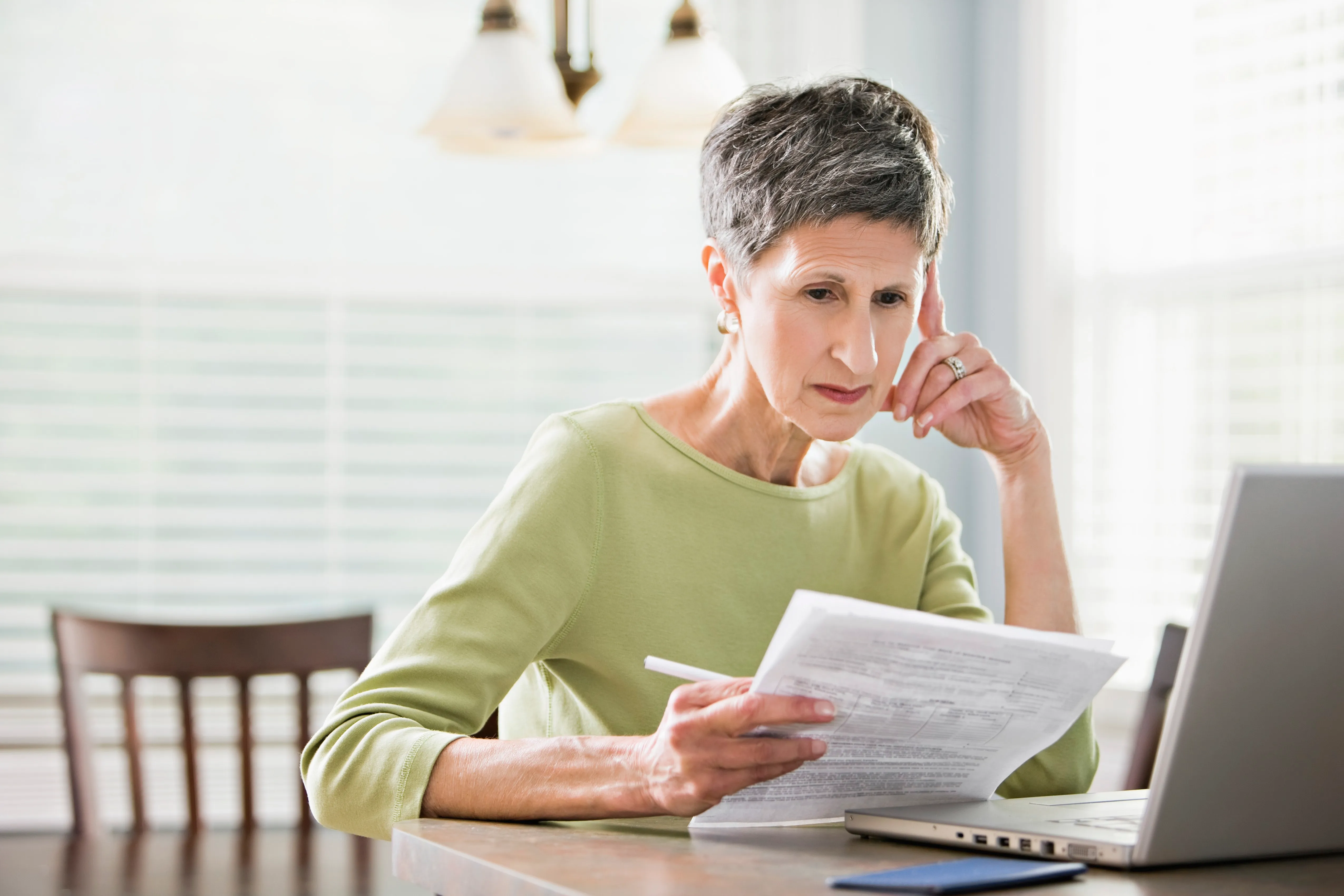 Senior woman reviewing documents while using a laptop.
