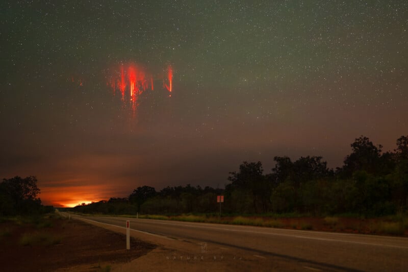 A dark, rural road stretches into the distance beneath a starry night sky. Above the horizon, bright red streaks known as sprites appear in the sky, while trees line both sides of the road.