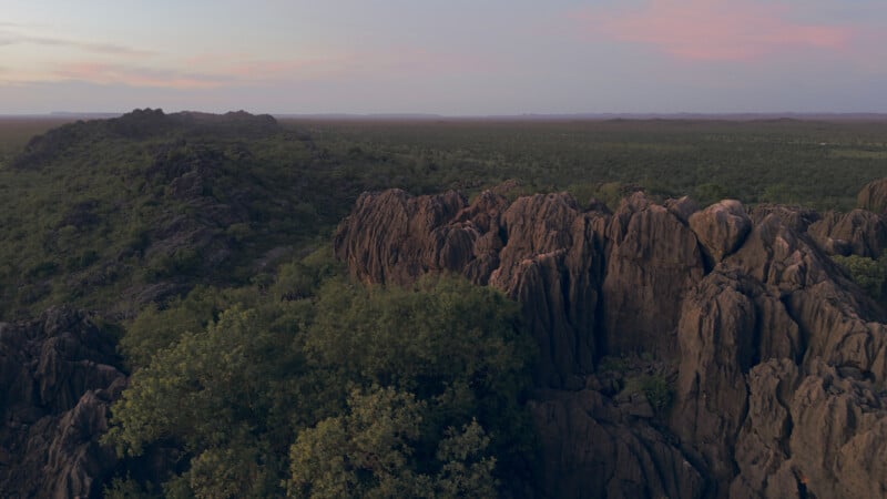 Aerial view of rugged rock formations surrounded by green vegetation, with a vast landscape and a pastel-colored sky at sunset or sunrise.