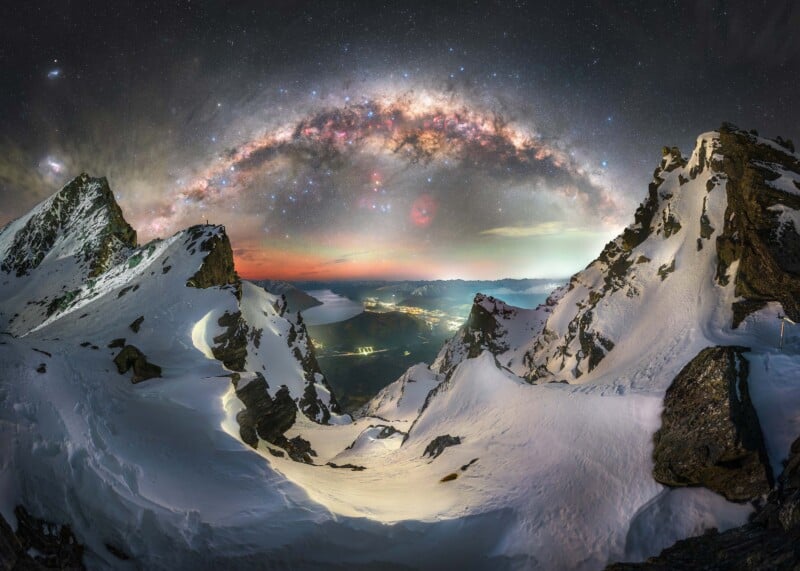 Snow-covered mountain peaks under a vibrant night sky, with the Milky Way arching above. In the distance, lights from a town glow warmly, contrasting with the dark, star-filled sky and rugged, icy terrain.