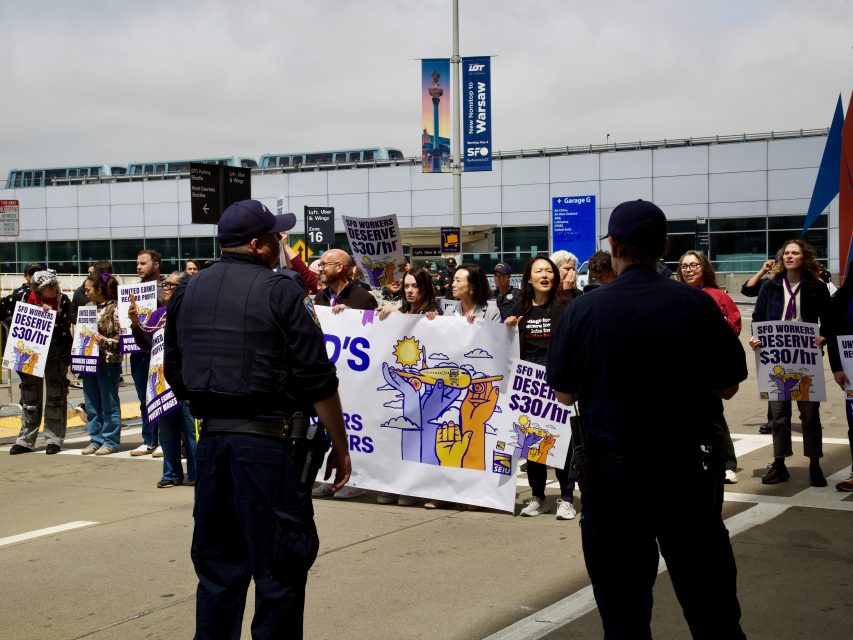 A group of protesters hold signs and banners demanding higher wages, as two uniformed officers stand nearby at an airport terminal, prepared to arrest anyone who disrupts the peace.
