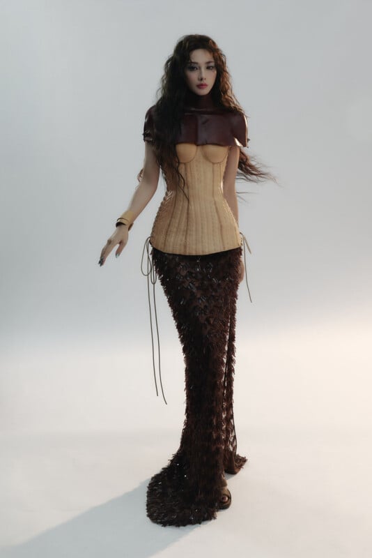 A woman stands against a plain background wearing a brown leather capelet, a beige corset, and a long, textured dark brown mermaid-style skirt. Her wavy hair is loose, and she has a neutral expression.