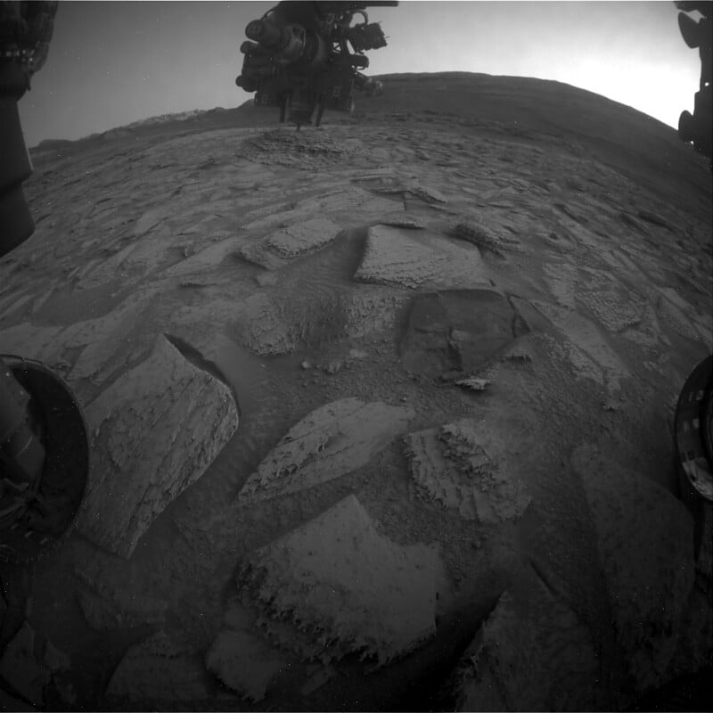 Black-and-white image from a Mars rover shows rocky, cracked terrain with square and irregular stones in the foreground; rover wheels and equipment are partially visible at the edges and top of the image.
