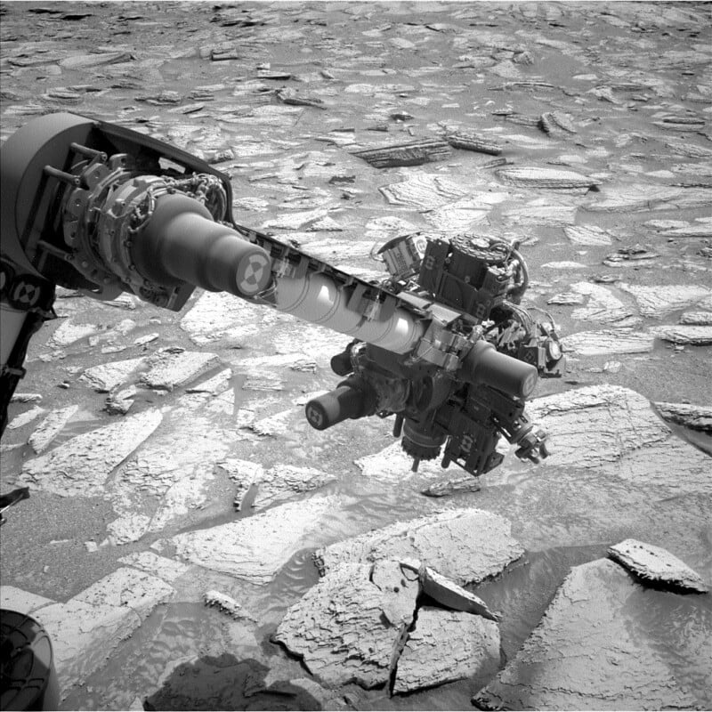 A robotic arm extends over a rocky, cracked surface on Mars, with scientific instruments visible at the arm’s end. The ground is covered in large, irregular stone slabs. The image is in black and white.