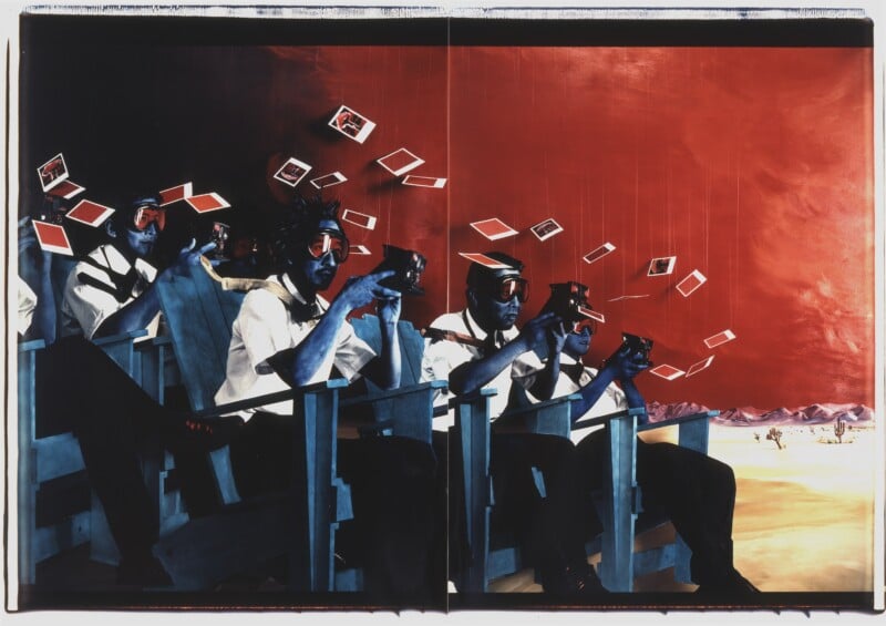 A row of people wearing goggles and holding cameras sit in bleacher seats, taking photos as snapshots float above them. The sky is a surreal red, and the desert landscape extends into the distance.