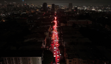 A long line of cars with bright red brake lights stretches down a dark city street at night, with illuminated buildings in the distant background.