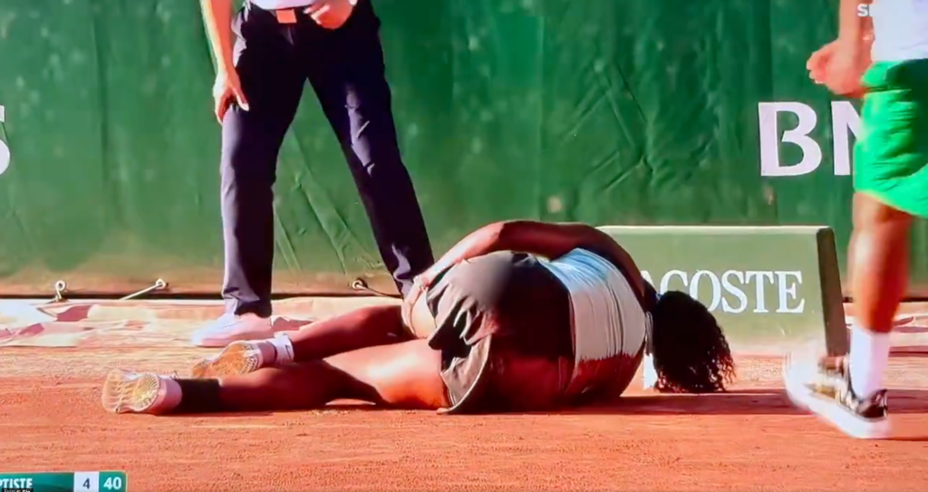 A tennis player in a black and white outfit is lying on the clay court, seemingly injured, while two officials stand over her.