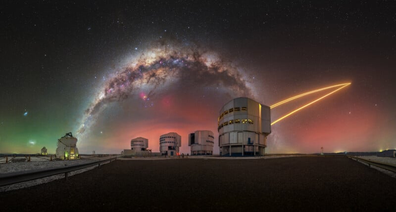 Five large observatory buildings stand under a starry night sky with the Milky Way arching above. A glowing streak of light curves upward, and green and red hues color the horizon.