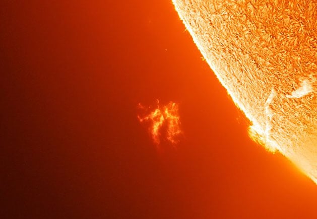 A close-up image of the sun’s surface shows a bright, textured edge with an orange solar prominence extending outward against the dark background of space.