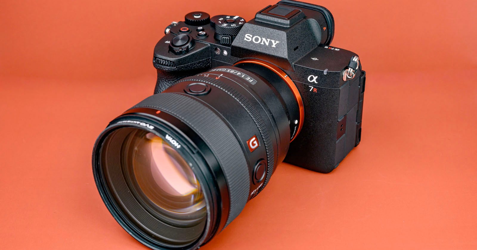 A Sony Alpha 7R mirrorless camera with a large zoom lens attached, placed on an orange surface with visible control dials and logos.