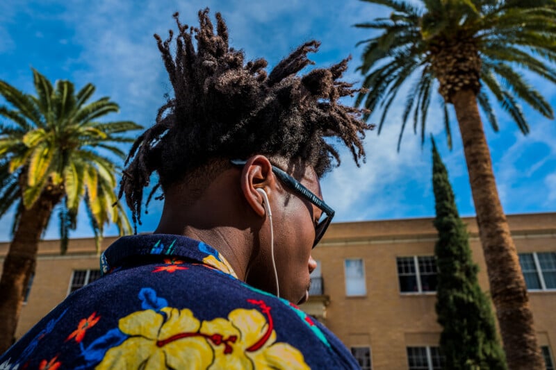 A person with dreadlocks and sunglasses listens to earphones while standing outdoors, wearing a blue floral shirt. Palm trees and a tan building with windows are in the background under a blue sky.