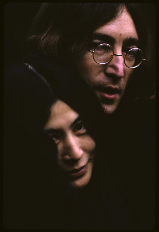 A close-up of two people with long dark hair. The person in front is partially smiling, while the person behind wears round eyeglasses and looks forward. Both are set against a dark background.