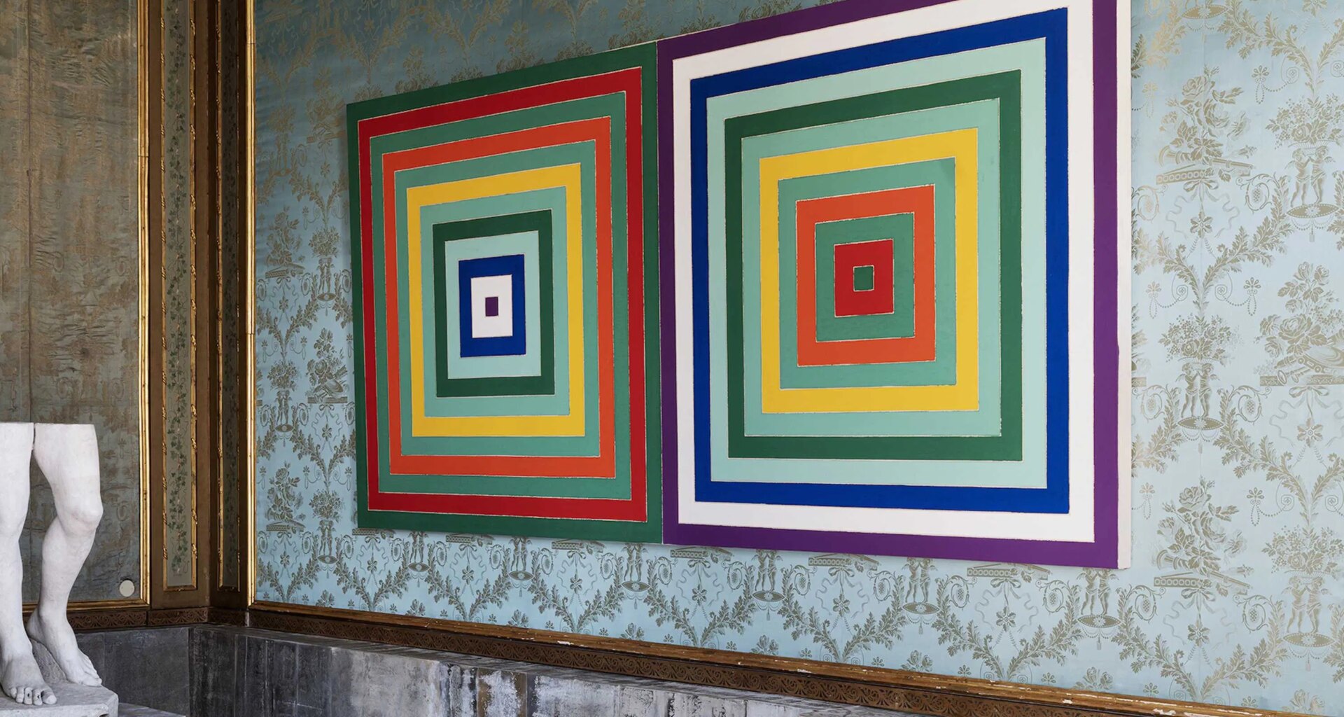 Colorful geometric artwork with concentric squares hangs on an ornate wall next to a partial view of a marble statue.