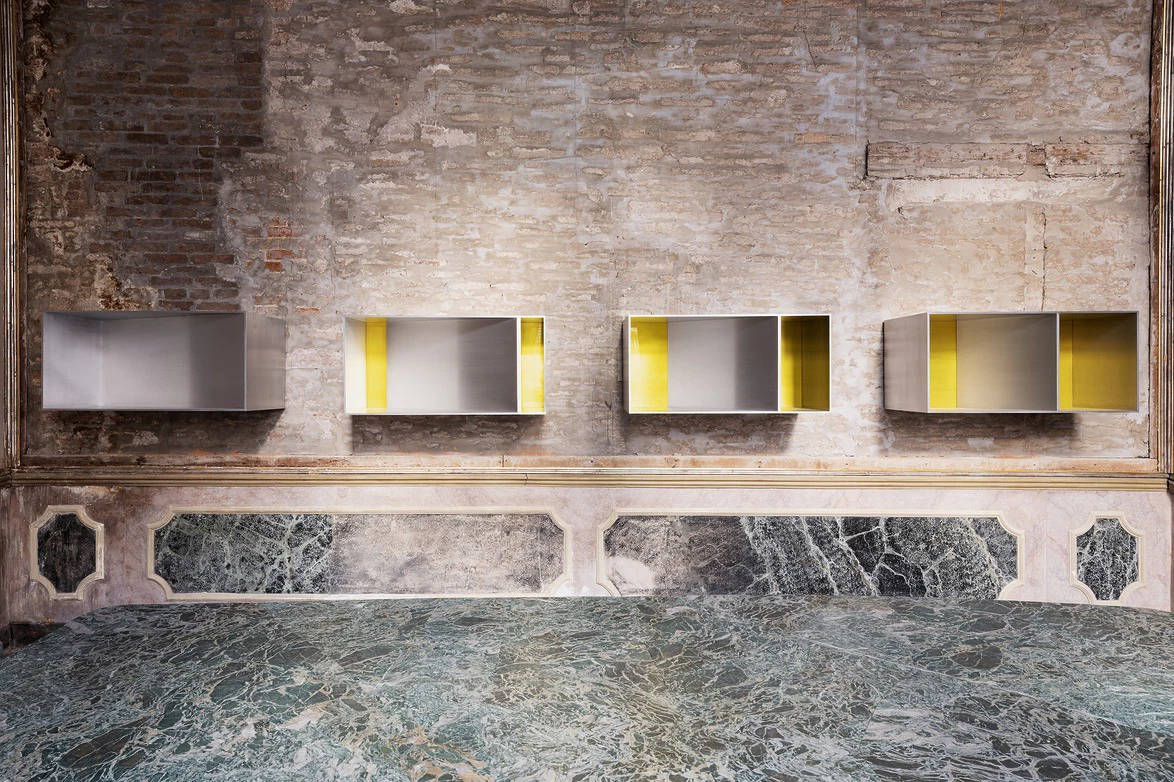 Four modern metal and yellow art installations on a rustic brick wall above a textured marble surface in a gallery setting.