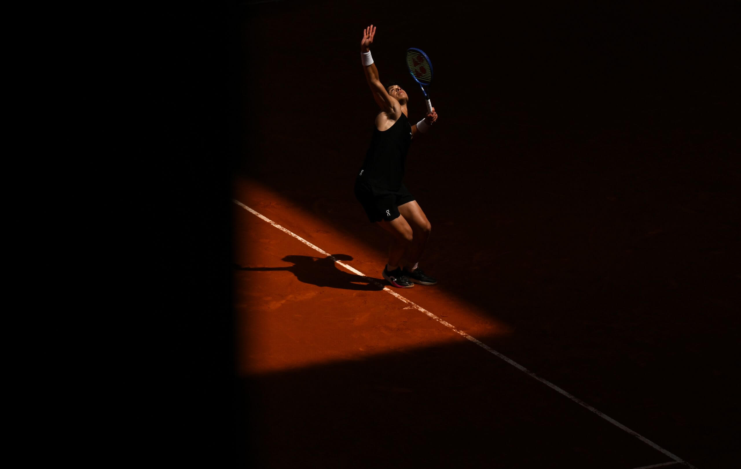 Ben Shelton hits a serve in a triangle of light on a clay court.