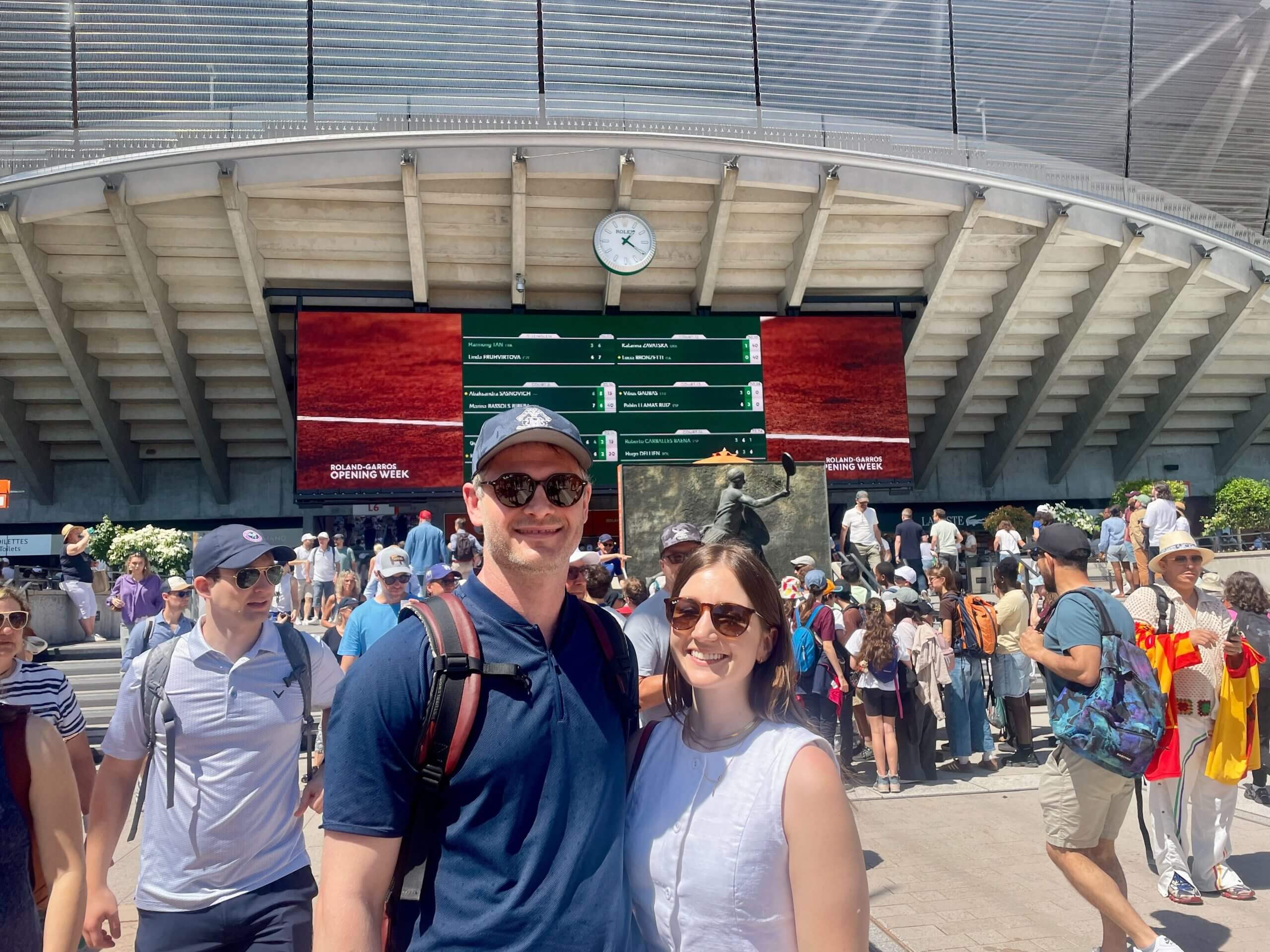 A man in a navy polo wears a baseball cap and a rucksack as he stands with a woman in a white top and sunglsses. They are both in front of a tennis stadium with a screen showing scores.