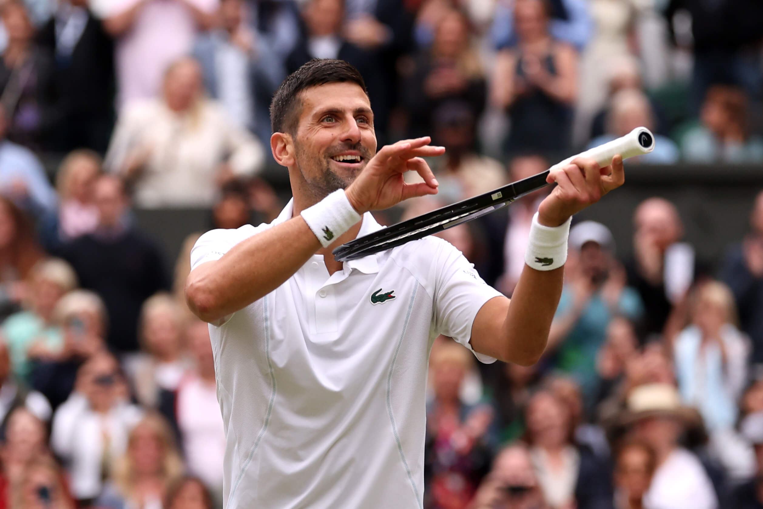Novak Djokovic pretends to play his tennis racket like a violin while wearing a white tennis outfit with a crowd in the background in soft focus.