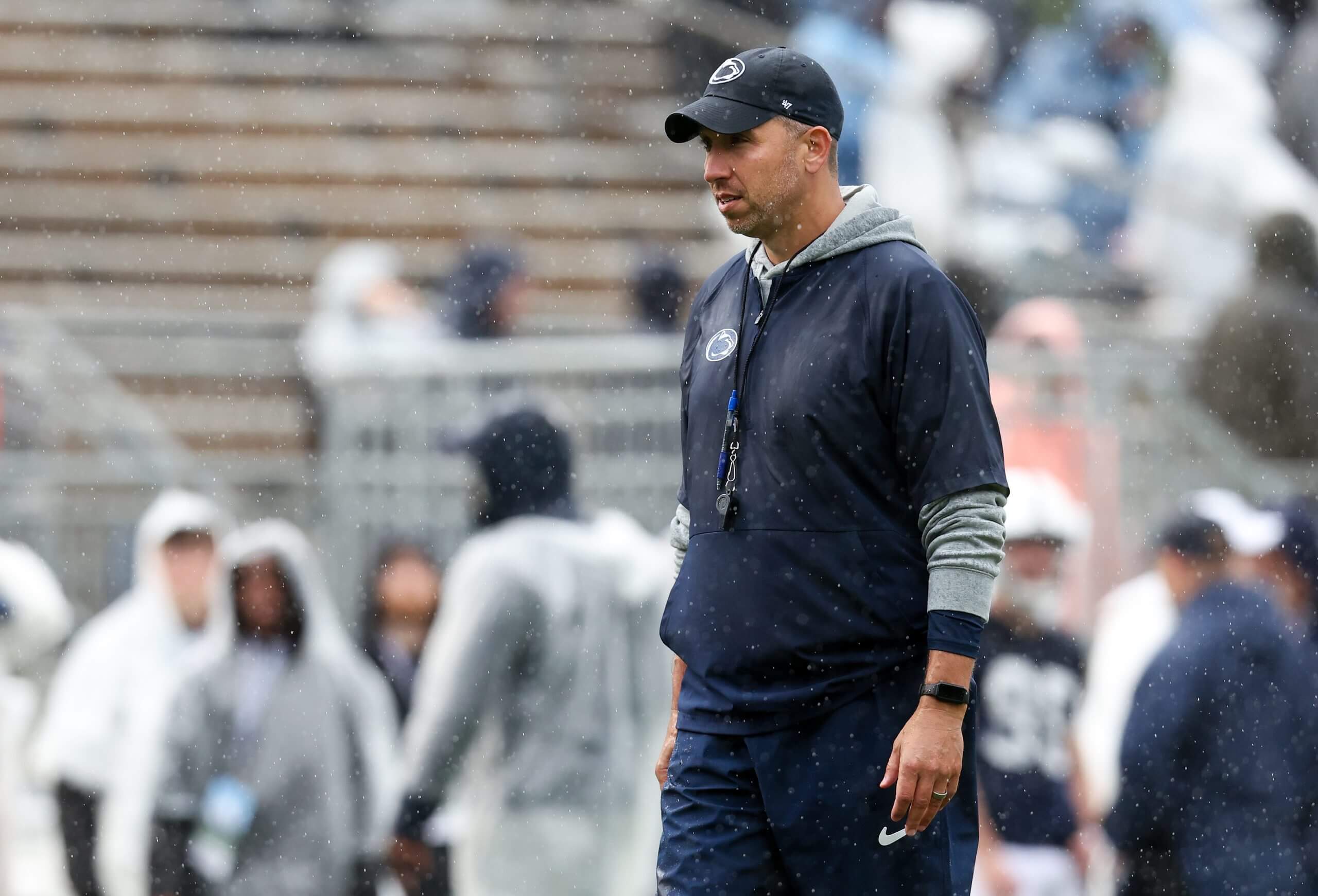 Matt Campbell walks on the field with a ball cap on a whistle around his neck with people and stadium seats blurred in the background.