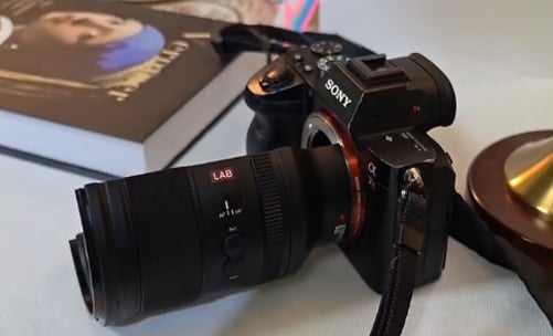 A Sony Alpha digital camera with a large lens is placed on a table next to a book featuring an artwork cover and another unidentified object.