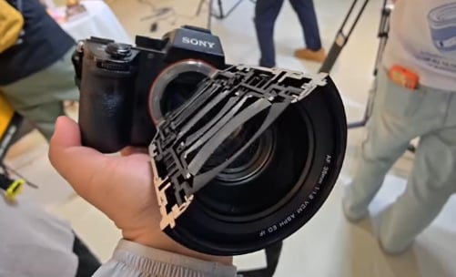 A person holds a Sony camera with a large lens. A metallic cut-out piece is partially covering the lens. People and camera equipment are visible in the blurred background.