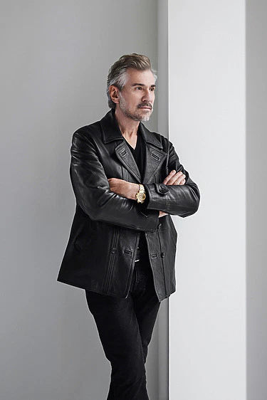 Man in a black leather jacket with arms crossed, standing by a white wall, looking thoughtful.