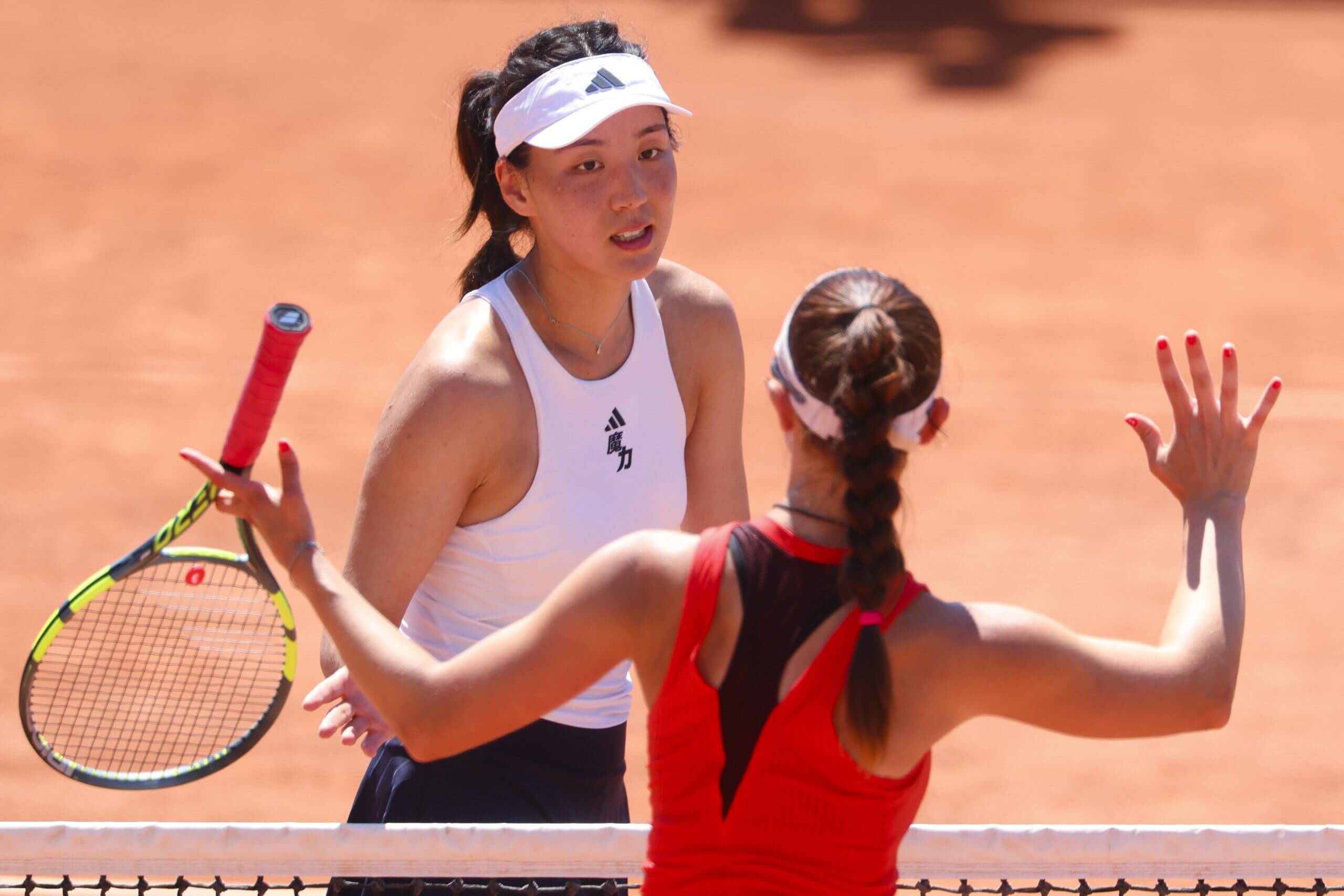 Wang Xinyu (left) and Tamara Korpatsch meet at a tennis net on a clay court with Korpatsch gesticulating with both arms.