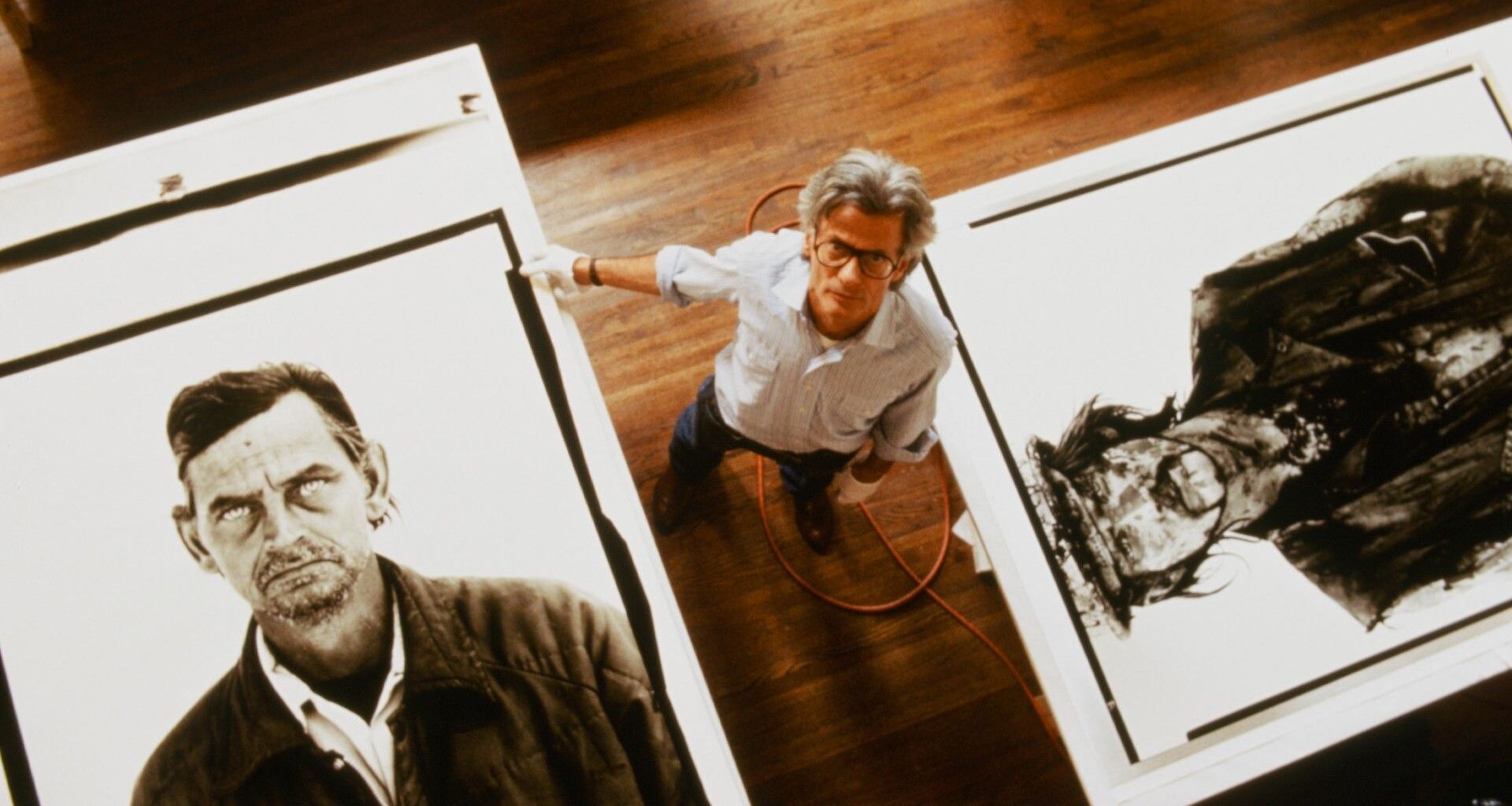 Shot from above, Avedon stands on a wooden studio floor between two enormous exhibition prints from his In the American West series, dwarfed by the faces around him.