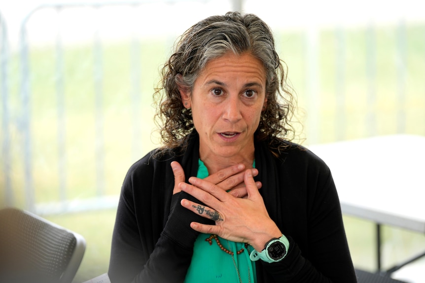 A woman with dark, greying hair speaking with her hands pressed to her chest
