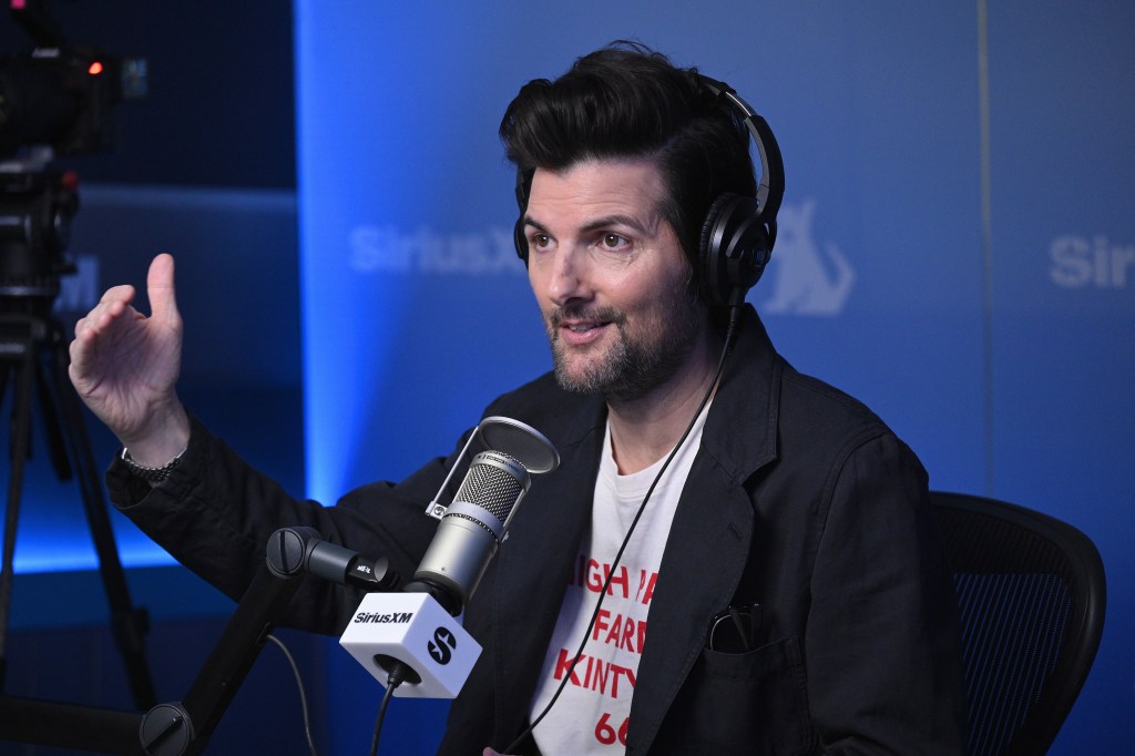 Adam Scott wearing headphones and a black jacket over a white t-shirt, speaking into a microphone at SiriusXM Studios.