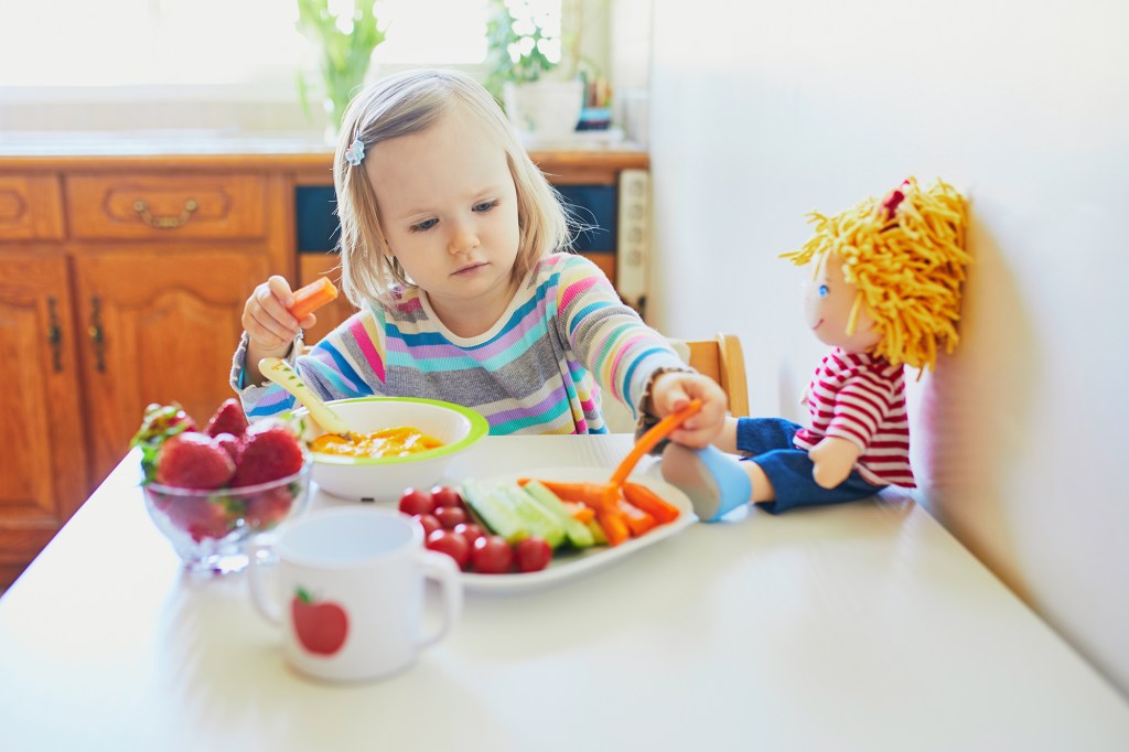 Toddler girl eating fresh fruits and vegetables while feeding her doll.