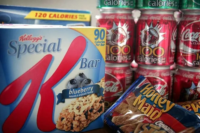 Special K blueberry bars, Coke cans, and Honey Maid graham crackers are shown, all emphasizing calorie content on their packaging