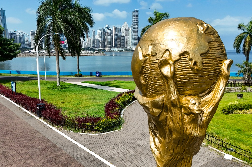 A giant replica of the FIFA World Cup trophy sits on a paved area with green grass and palm trees, overlooking a bay with a city skyline in the background.