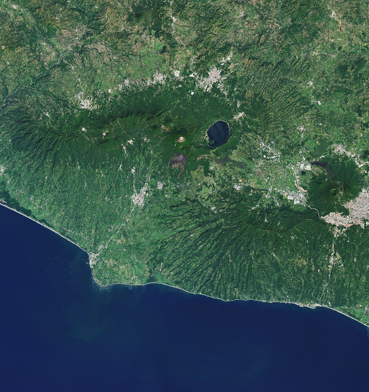 A chain of forested volcanic peaks arc across the image, with an area of dark lava flows in the lower right and the brighter urban area of Ahuachapán in the upper left.