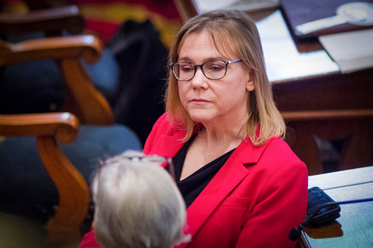 A person wearing glasses and a red blazer is seated, looking towards someone else in the foreground. There is a wooden chair and papers on a desk in the background.