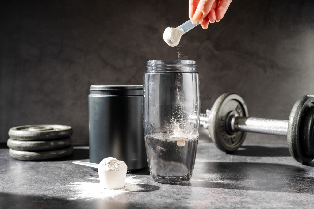 A hand pouring creatine powder from a scoop into a water bottle next to a container of supplements and a dumbbell.