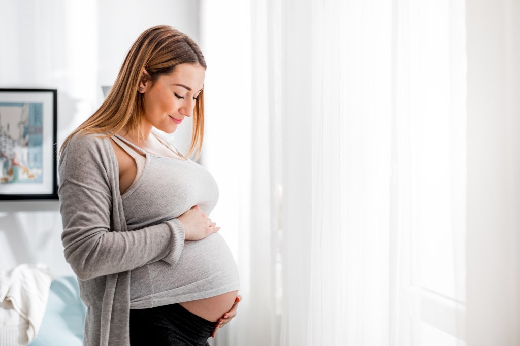 A pregnant woman in a gray top and cardigan, gently touching her belly while standing by a window, smiling down at her stomach.