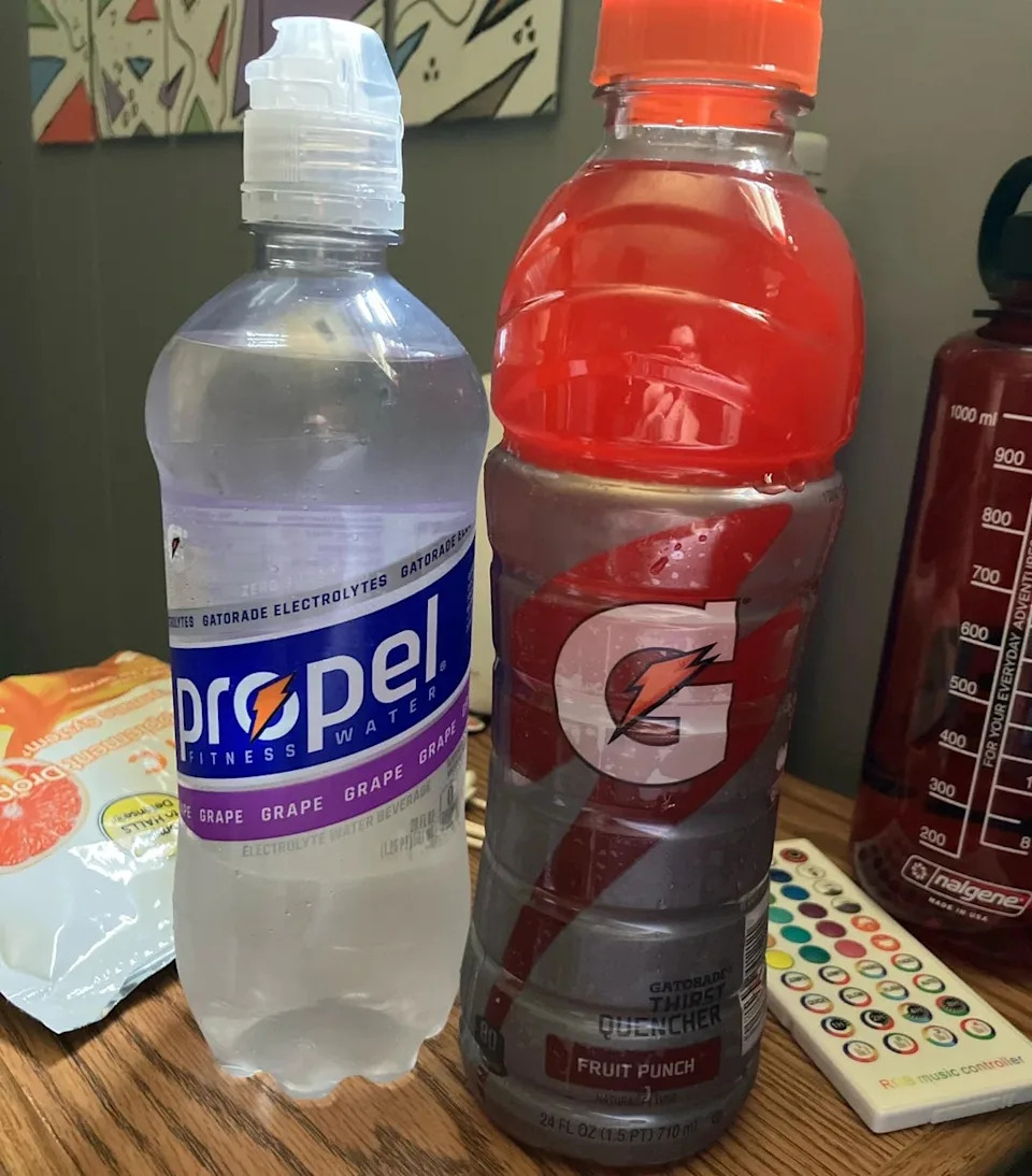 Two sports drinks on a table: a Propel fitness water bottle and a Gatorade Fruit Punch bottle. In the background, a large water bottle and snacks
