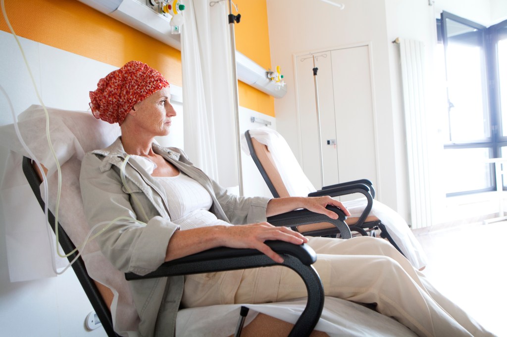A female cancer patient, wearing a red headscarf, receiving outpatient chemotherapy treatment through an IV drip while sitting in a hospital room.